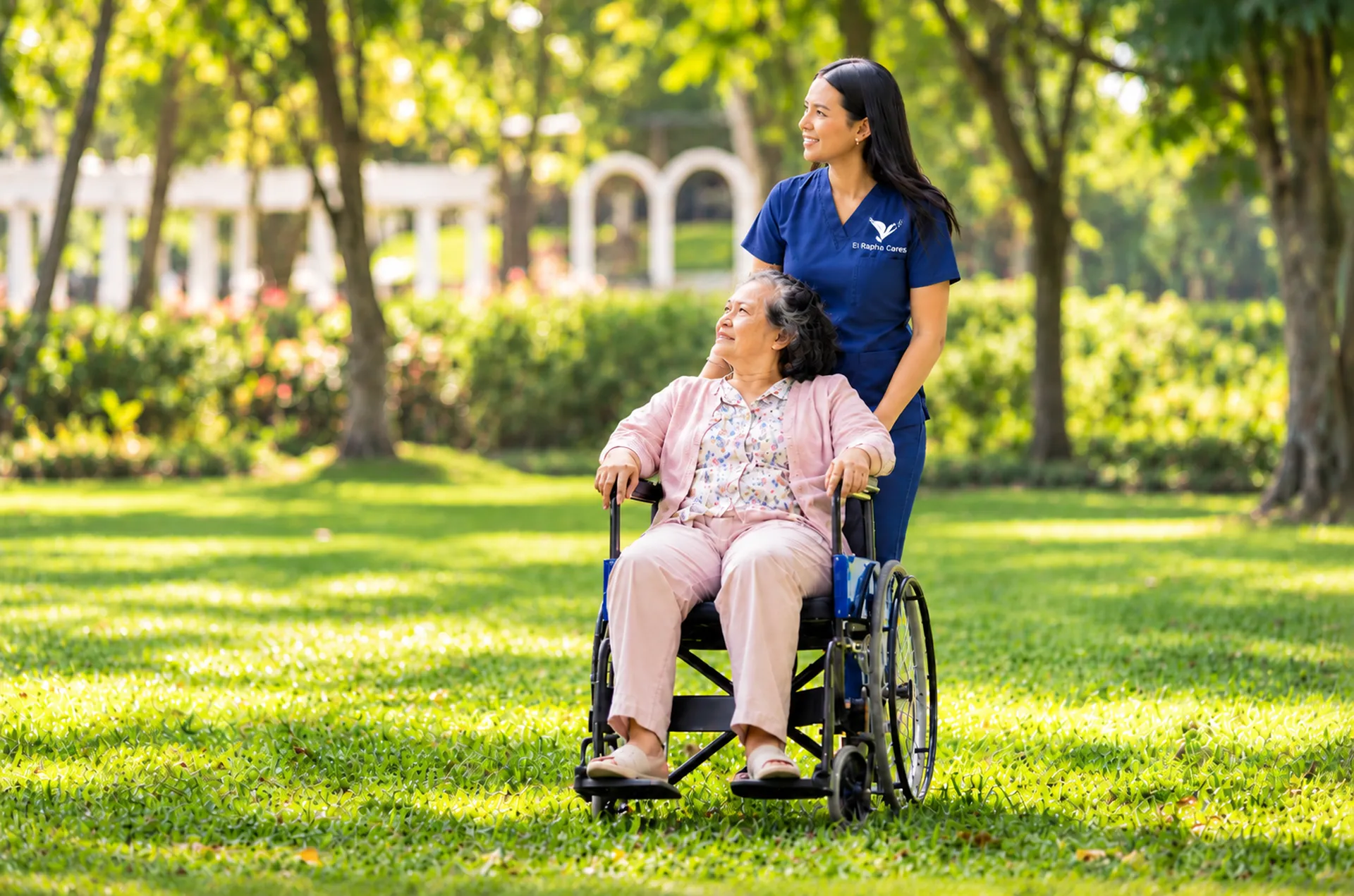 Carer pushing an elderly woman in a wheelchair through a sunny park